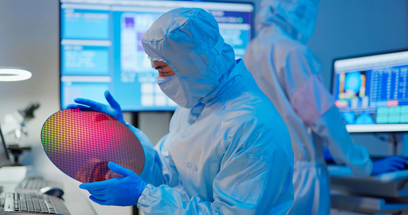 Técnico de semiconductores con equipo completo de protección para salas limpias y guantes azules inspeccionando una oblea de silicio reflectante, con pantallas de monitoreo digital y otro técnico trabajando en segundo plano.